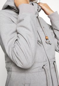 Gray hooded jacket with a textured finish, featuring a braided drawstring with wooden accents and elastic cuffs. Zippered front pockets included.