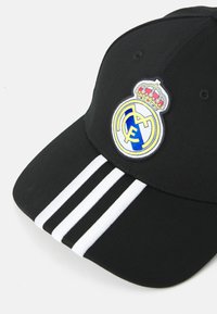 Casquette noire avec une visière courbée, comportant un logo Adidas blanc brodé et trois rayures blanches. Inclut un écusson rond brodé du Real Madrid.