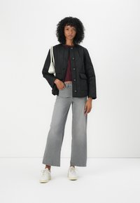 Vrouw met krullend haar die een zwarte gewatteerde jas, een bordeauxrood topje, een grijze brede spijkerbroek, witte sneakers draagt en een kleine witte schoudertas bij zich heeft.