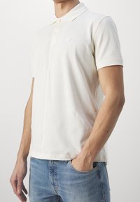 Krämfärgad polo skjorta i bomull, med klassisk krage, tvåknappslå, korta ärmar och ett litet emblem på bröstet.