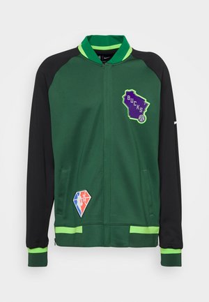 Veste verte et noire avec une fermeture éclair complète, affichant un logo "BUCKS" violet et des écussons en forme de losange avec des accents rouges, bleus et blancs.