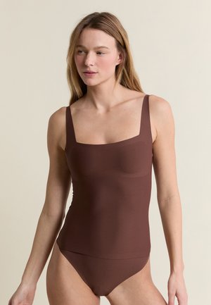 Młoda kobieta o długich blond włosach, ubrana w brązowe, bezrękawnikowe body z kwadratowym dekoltem, stojąca na beżowym tle.