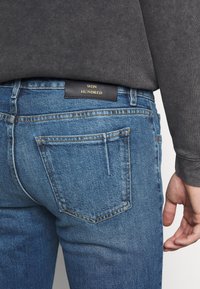 Los vaqueros de mezclilla azul presentan un corte clásico con un diseño limpio, costuras visibles y una etiqueta negra que dice "WON HUNDRED" en la cintura.