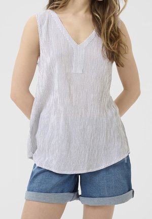 Woman wearing a sleeveless white V-neck top with thin vertical stripes and rolled-up blue denim shorts against a neutral background.