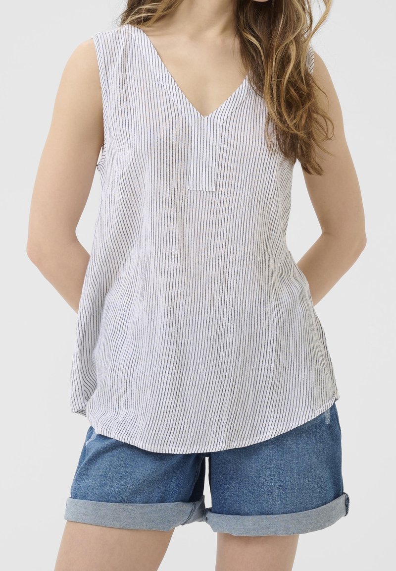 Femme portant un haut blanc sans manches à col en V avec de fines rayures verticales et un short en jean bleu retroussé sur un fond neutre.