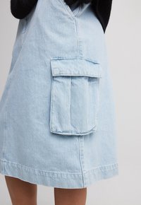 Abito in denim azzurro chiaro con una grande tasca frontale, dettagli cuciti e un taglio semplice e dritto. Il tessuto appare morbido e strutturato.