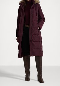 Burgundská dlouhá péřová bunda s kožíškem uvnitř kapuce, se zapínáním na zip a patentky, s bočními kapsami, v kombinaci s černými kalhotami a hnědými vysokými kozačkami.