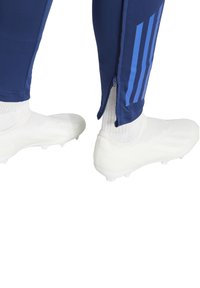 Marineblaue Sporthosen mit blauen Streifen und Reißverschlüssen an den Knöcheln, kombiniert mit weißen Schläppchen und gerippten weißen Socken.
