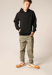 Zwarte hoodie met textuur en een buidelzak, gecombineerd met licht kaki cargobroek met zijritsen. Witte en oranje sneakers maken de outfit compleet.