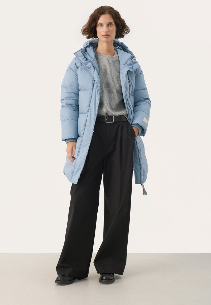 Helle blaue Steppjacke mit Kapuze, ausgestattet mit einem Reißverschluss, großen Taschen und einem längeren Schnitt, kombiniert mit einem grauen Pullover und schwarzen weit geschnittenen Hosen.