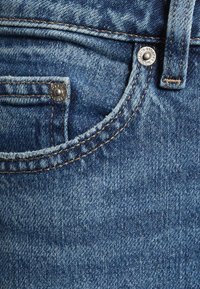 Primer plano del bolsillo de unos vaqueros de mezclilla azul con costuras visibles, remaches y detalle de la trabilla del cinturón.