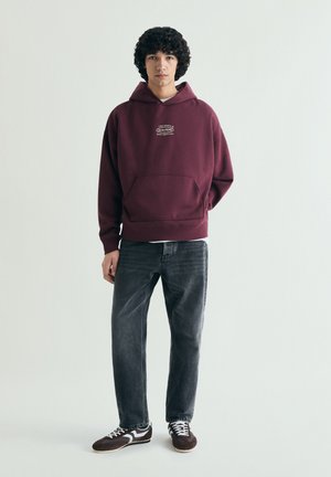 Tânăr cu păr creț, purtând un hanorac bordo cu buzunar frontal, blugi negri și adidași negri cu alb, stând în picioare pe un fundal simplu.