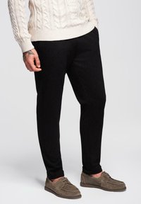 Homme portant un pantalon noir, un pull en tricot crème à motif torsadé et des chaussures à lacets en daim marron, se tenant devant un fond clair.