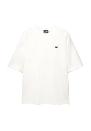 Valkoinen oversized-t-paita, valmistettu puuvillasta, jossa on lyhyet hihat ja pieni punainen logo rinnassa. Pyöreä kaula-aukko, minimaalinen muotoilu.
