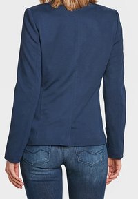 Persona vestida con una chaqueta azul marino ajustada y jeans de mezclilla azules, vista desde atrás contra un fondo claro y liso.