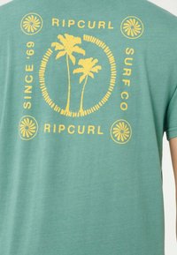 Maglietta verde con grafica di una palma gialla, testo "Ripcurl", "Since '69" e "Surf Co" intorno al disegno, con accenti floreali a cerchio.
