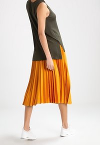 Olivengrøn ærmeløs top kombineret med et plisseret, knælængde orange nederdel. Hvide sneakers fuldender outfittet.