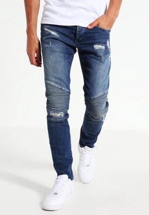 Homme portant un jean slim bleu délavé avec des panneaux côtelés aux genoux et des baskets blanches, marchant avec une main dans la poche.