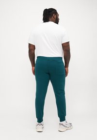 Teal joggings met een taps toelopende pijp, voorzien van een achterzak en geribbelde manchetten, gecombineerd met een wit shirt met korte mouwen.