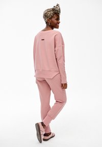 Kobieta uśmiechająca się i patrząca za siebie, ubrana w różową bluzę i spodnie, różowe trampki oraz panterkową chustę na głowie na białym tle.