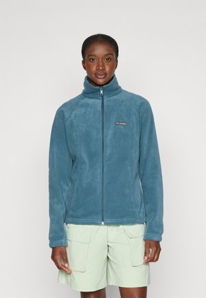 Veste en polaire teal avec un col haut, fermeture éclair intégrale et logo sur la poitrine gauche ; portée avec un short vert clair. Texture douce et design ajusté.