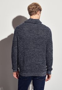 Mörkblå stickad tröja med sjalkrage och långa ärmar. Texturerad tyg med ett subtilt mönster, som slutar vid höfterna. Bärs med mörka jeans.