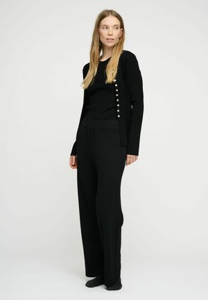 Musta neulottu neuletakki, jossa helmipainikkeet, yhdistettynä mustaan toppiin ja leveälahkeisiin housuihin; kaikissa on sileä pinta ja rento istuvuus.