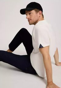 Biała koszula z krótkim rękawem z granatowym trójkątnym akcentem na rękawie, połączona z ciemnoniebieskimi dżinsami. Czapka z czarnej kordury dopełnia strój.