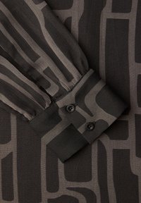 Rękaw bluzki w czarno-szary wzór z mankietem ozdobionym dwoma czarnymi guzikami. Tkanina ma gładką, przejrzystą teksturę z pionowymi paskami.