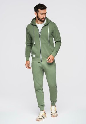 Uomo con felpa con cappuccio color verde oliva con zip e pantaloni da jogging abbinati, che guarda verso il basso, in piedi davanti a uno sfondo semplice di colore grigio chiaro.