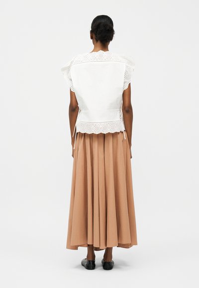 Γυναίκα με μαύρα μαλλιά σε κότσο, που φοράει λευκό πουκάμισο από δαντέλα eyelet και μακριά μπεζ πλισέ φούστα, στέκεται με την πλάτη, φορώντας μαύρα φλατ παπούτσια.