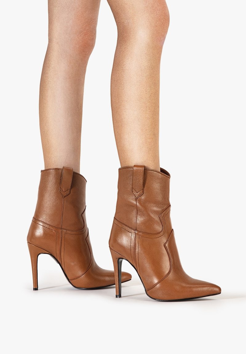 Jambes portant des bottes en cuir marron à talons hauts, hauteur mi-mollet, avec bouts pointus et languettes de tirage, sur fond blanc.