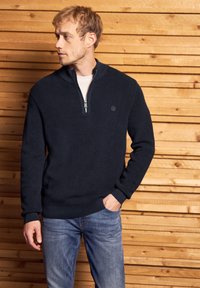 Pull en maille marine avec un col zippé à mi-hauteur et une texture côtelée. Présente un petit logo sur la poitrine ainsi que des poignets et un ourlet ajustés. Jeans portés en dessous.