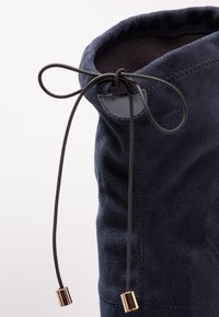 Bota de ante azul marino con textura suave, que presenta un cierre de cordón con extremos acentuados en cobre y un interior suave.