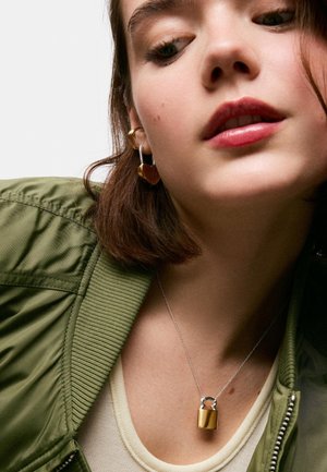 Młoda kobieta w zielonej kurtce, kremowym topie, złote kolczyki w kształcie serca i złoty naszyjnik z zawieszką w kształcie kłódki.