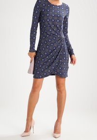 Long-sleeve navy dress with circular patterns, wrap design, and textured fabric. Paired with light pink high heels for contrast.