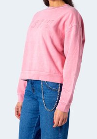 Femme portant un sweat-shirt Levi's rose et un jean bleu avec une chaîne argentée attachée à la boucle de ceinture avant, la main reposant sur le côté.
