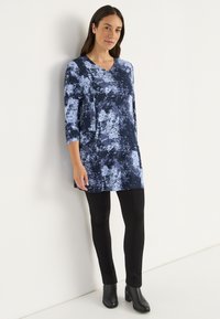 Marineblaue Tunika mit einem Spritzmuster in Hellblau, mit V-Ausschnitt und drei Viertelärmeln, kombiniert mit schwarzen, figurbetonten Hosen und Stiefeletten.