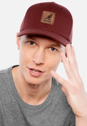 Giovane uomo dalla pelle chiara che indossa un cappellino Kangol color melanzana e una maglietta grigia, alzando la mano vicino al volto su uno sfondo uniforme.