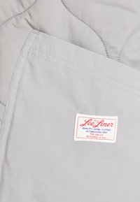 Tissu matelassé gris à la texture lisse. Présente une étiquette rouge et bleue portant la mention "Lee Liner Quality Work Clothes" sur un fond blanc.