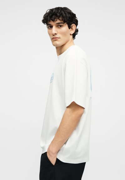 Νεαρός άνδρας με καστανά σγουρά μαλλιά, φοράει φαρδιά λευκή μπλούζα και μαύρο παντελόνι, στέκεται πλαγιαστά με τα χέρια στις τσέπες σε λευκό φόντο.