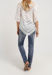 Femme vue de dos, portant un haut en crochet blanc, un jean bleu et des sandales, se tenant devant un fond uni.