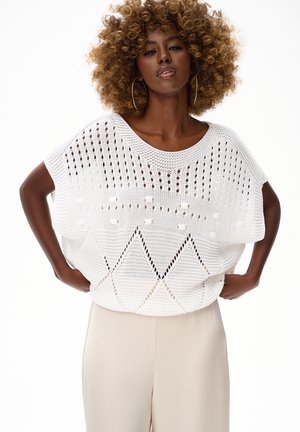 Kobieta o kręconych włosach, nosząca duże kolczyki koła oraz biały, luźny, dziany top z geometrycznymi wzorami, zestawiony z beżowymi spodniami z wysokim stanem.