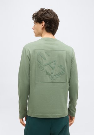 Hombre con cabello corto y ondulado que lleva una camisa de manga larga verde claro con el logo en relieve "EA7 Emporio Armani" en la espalda, de espaldas.