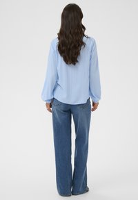 Frau mit langen braunen Haaren, die eine hellblaue Bluse und blaue Jeans trägt, steht mit dem Rücken zu einem schlichten Hintergrund.