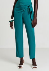 Pantalons taille haute en bleu sarcelle avec détail en tissu noué, portés avec des chaussures à talons ouverts blanches et des bracelets empilés au poignet gauche.