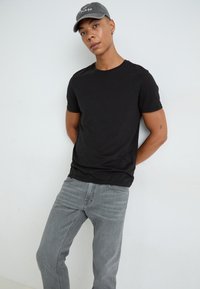 Camiseta negra de manga corta con cuello redondo, combinada con jeans gris claro. El modelo lleva una gorra gris con texto bordado.