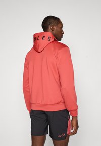 Haut à capuche rouge avec imprimé noir "ENDLESS" sur la capuche, présentant une texture lisse, un design ajusté et des poignets côtelés, associé à des shorts sombres.