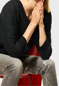 Svart stickad tröja med V-hals och trekvartslånga ärmar, i kombination med blanka, texturerade silvriga jeans. Röd stol i bakgrunden.