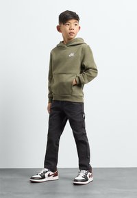 Olīvu zaļa kapucēta džemperis ar kangaroo kabatu un baltu Nike logo, apvienots ar melnām kravas biksēm un melnajiem/sarknajiem sporta apaviem ar baltām akcentiem.